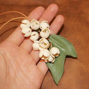 May include: A handcrafted necklace featuring a cluster of off-white flower pendants and two green leaves. The pendant is suspended from a thin, brown leather cord. The necklace is held in a hand.