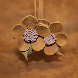 Può includere: Due fiori di cuoio marrone con centri rosa. I fiori sono attaccati a un cordoncino di cuoio marrone.