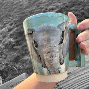 May include: Hand-painted ceramic mug featuring an elephant design. The mug has a light blue rim and handle, with a gray and white elephant face on the front. The mug is held by a person.