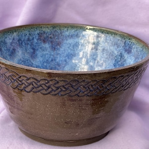 May include: A handmade ceramic bowl with a brown exterior and a blue interior. The rim features a dark blue Celtic knot design. The bowl is likely used for serving food or as a decorative piece.