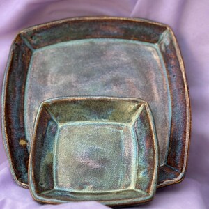 May include: Two square ceramic serving dishes with rounded corners. The dishes have a teal and brown glaze, with a metallic sheen. The larger dish is behind the smaller one, both resting on a lavender fabric.