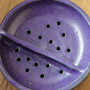 May include: A round, purple ceramic soap dish with a glossy, speckled finish. The dish features a central divider that splits the interior into two sections, each containing multiple drainage holes to allow water to pass through the base.