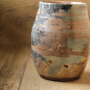 May include: A ceramic vase with a rounded body and a wide opening. The surface features a mottled finish with blended tones of terracotta, charcoal black, and muted sage green, creating a weathered appearance with visible texture and color variations.