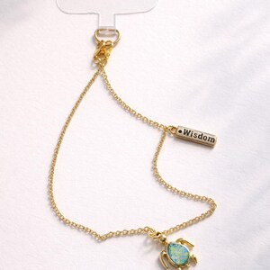 May include: Gold-colored phone charm with a turtle pendant and a small rectangular charm that reads "Wisdom." The turtle pendant has a blue-green opal-like center. The charm has a clear plastic loop for attachment.