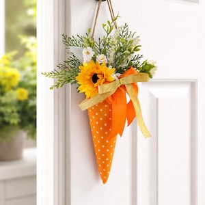 May include: A door hanging decoration. It features an orange, cone-shaped fabric container with white polka dots, filled with greenery, white flowers, and a yellow sunflower. A gold ribbon and bow complete the design.