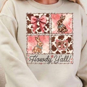 May include: Beige sweatshirt with a four-panel design featuring a pink bow, a bunny, a pretzel, and a bunny. The design incorporates cow print and floral elements. The text "Howdy Y'all!" is written in a script font below the panels.