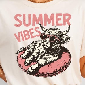 Peut inclure: Un t-shirt à manches courtes de couleur crème avec une illustration d'une vache Highland portant des lunettes de soleil roses sur une bouée rose. Le texte "SUMMER VIBES" est imprimé en rose au-dessus de l'illustration dans une police usée.