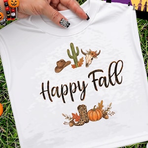 Puede incluir: Una camiseta blanca sin mangas con un estampado gráfico que incluye el texto Happy Fall en letra cursiva marrón. El diseño presenta un sombrero de vaquero marrón, un cactus verde, una calavera de vaca, una bota de leopardo y una calabaza.