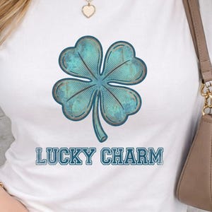 May include: White t-shirt featuring a large, distressed teal four-leaf clover graphic. The text "LUCKY CHARM" is printed below the clover in a matching teal, vintage-style font. The shirt is a casual style.
