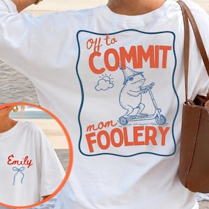 May include: A white long-sleeve shirt featuring a large back graphic of a frog wearing a hat on a scooter with the text "off to COMMIT mom FOOLERY" and a small front chest graphic with the name "Emily" in red script above a blue line-art bow.