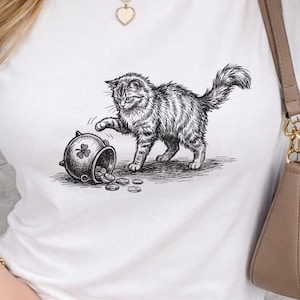 Puede incluir: Camiseta blanca con una ilustración en blanco y negro de un gato y una olla de oro. El gato está jugando con la olla volcada, con monedas esparcidas. El diseño es detallado y caprichoso.