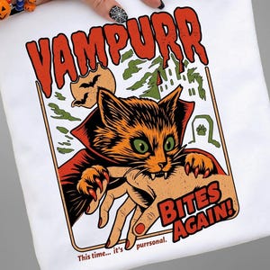 Puede incluir: Una camiseta blanca con un gráfico de un gato naranja vestido de vampiro mordiendo una mano humana. El texto dice VAMPURR BITES AGAIN! y This time... it's purrsonal. El fondo muestra una luna, murciélagos y la silueta de un castillo.