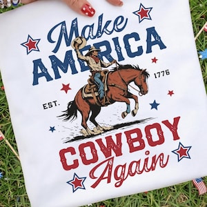 Puede incluir: Una camiseta blanca con un estampado gráfico de un vaquero montando un caballo marrón. El texto "Make America Cowboy Again" aparece en azul y rojo, con "EST. 1776" impreso cerca. El diseño incluye estrellas rojas y azules alrededor.