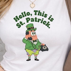 May include: White t-shirt with a cartoon graphic of a character dressed as a leprechaun, holding a phone. The text "Hello, This is St. Patrick" is arched above the character in green letters.