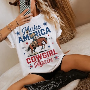 Puede incluir: Una camiseta blanca de manga corta con un estampado gráfico que incluye el texto "Make America COWGIRL Again" en azul y rojo. El diseño muestra a una vaquera montando un caballo marrón, el año 1776 y varios detalles de estrellas.