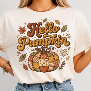 Peut inclure: Un t-shirt à manches courtes de couleur crème avec une impression graphique portant le texte Hello Pumpkin en lettres cursives marron et jaune. Sous le texte se trouve une grosse citrouille au motif patchwork floral et rayé aux tons automnaux.