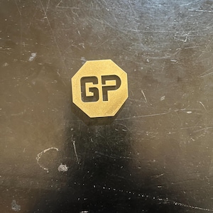 May include: A gold-colored, octagonal metal object with the letters "GP" cut out in the center. The object is set against a dark, textured surface.