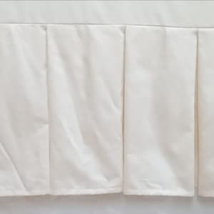 May include: Cream-coloured bed skirt with a pleated design. The fabric is smooth and appears to be satin or similar material. The pleats are evenly spaced and run the length of the skirt, creating a tailored look. The skirt is designed to fit around the base of a bed.