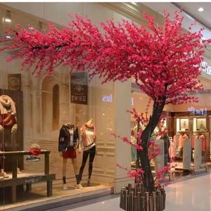 Può includere: Un vibrante albero artificiale rosa con un tronco scuro e una base decorativa si erge di fronte a una vetrina. I rami dell'albero sono carichi di fiori rosa, aggiungendo un tocco di colore alla scena. I manichini sono visibili all'interno del negozio.