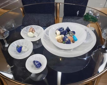 May include: Four white selenite bowls of varying sizes, with the largest bowl containing a collection of colorful gemstones, including blue, green, and orange stones. The bowls are arranged on a black glass table.