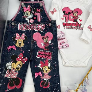 May include: A denim overall and a white long-sleeve bodysuit set. The overall features Minnie Mouse graphics, the name "Kensley", and pink accents. The bodysuit has a Minnie Mouse graphic and the text "Kensley's 1st Birthday Girl!"