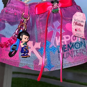 May include: Pink denim skirt with cross and heart designs, embellished with rhinestones. Features a pink bow, a charm with a cartoon figure, and the text "Demon Hunters". Includes a tag that says "Sewn by Shatina".