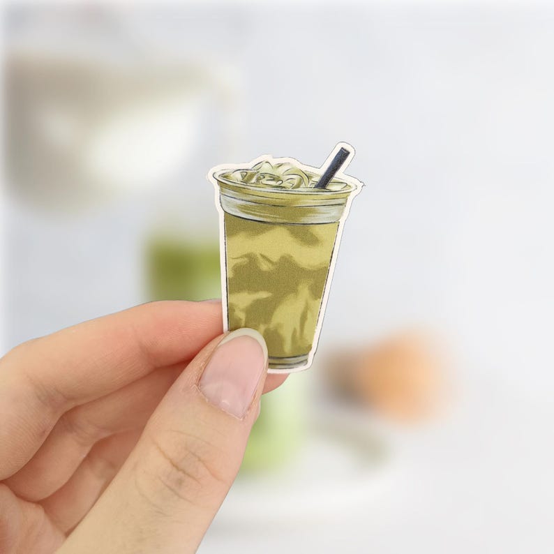 May include: A sticker of a matcha green tea drink in a clear plastic cup with a black straw. The drink has a frothy top and a light green color. The sticker is held by a person's hand.