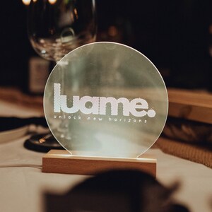 May include: A round, illuminated sign with the word "luame." in white, and the phrase "unlock new horizons" below it. The sign is mounted on a rectangular wooden base. The background is dark, with a wine glass visible.