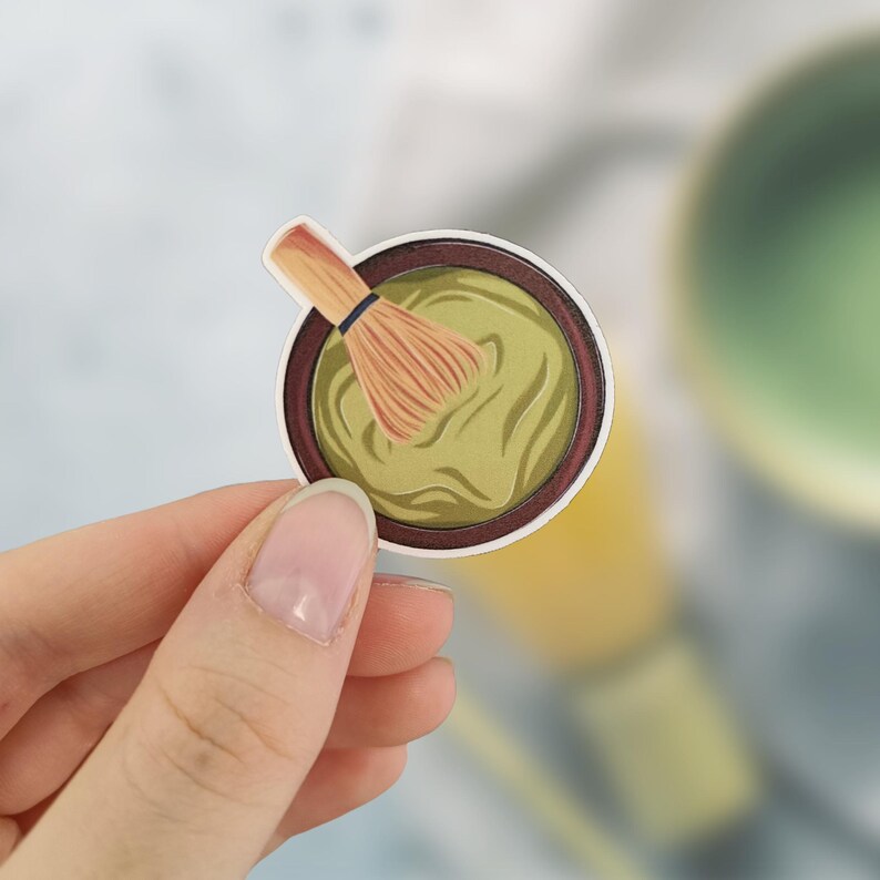 May include: A sticker featuring a matcha bowl with a bamboo whisk. The bowl is filled with green matcha and has a dark brown rim. The whisk is light brown with a black band. The sticker is held by a hand.