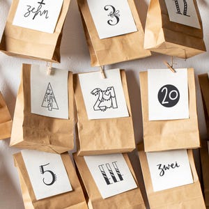 Könnte beinhalten: Adventskalender mit braunen Papiertüten, jede mit einer Zahl oder einem Wort auf einer weißen Karte. Die Tüten hängen an einer Schnur mit kleinen Holzklammern, was eine festliche Dekoration ergibt.