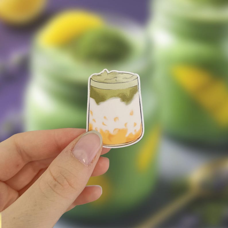 May include: A sticker of a layered drink with a green top, white middle, and orange bottom. The sticker is held in a person's hand. The background is blurred, showing a similar drink and a purple surface.