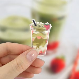 May include: A sticker of a matcha strawberry drink in a clear plastic cup. The drink has layers of green matcha, white cream, and pink strawberry syrup. A black straw and a strawberry slice garnish the top.
