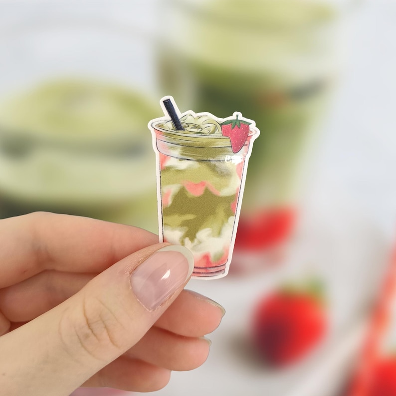 May include: A sticker of a matcha strawberry drink in a clear plastic cup. The drink has layers of green matcha, white cream, and pink strawberry syrup. A black straw and a strawberry slice garnish the top.