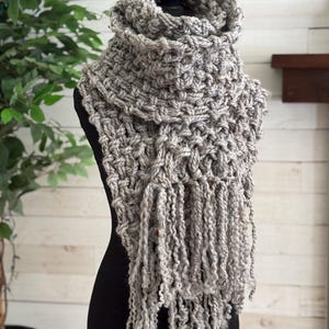 May include: A chunky knit scarf in a light gray heathered yarn featuring a thick, textured weave and long, loose fringe detailing at the bottom. The scarf is wrapped around a black mannequin, displaying a dense, looped stitch pattern throughout.