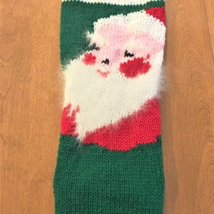 May include: A knitted Christmas stocking with a Santa Claus design. The stocking has a white top with the name "JOHN" in black letters, a green body, and a red and white Santa face. The Santa face has a pink face, red cheeks, and a white beard.