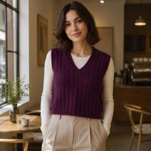 Könnte beinhalten: Eine dunkelviolette ärmellose Strickweste mit gerippter Textur und V-Ausschnitt. Das Kleidungsstück wird über einem cremefarbenen Langarmshirt getragen und mit einer hellbeigen Hose kombiniert, was einen Lagen-Look mit klarer Silhouette ergibt.