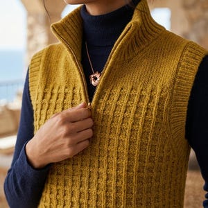 May include: Mustard yellow knitted vest with a high collar and a gold zip. The vest features a textured, vertical pattern. A gold necklace with a pendant is visible. The vest is worn over a navy blue turtleneck.