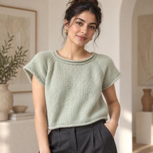Puede incluir: Un suéter de punto de manga corta en color verde salvia con textura acanalada, cuello redondo con borde enrollado y mangas raglán. El suéter tiene un corte corto y se combina con pantalones de color gris carbón oscuro con cintura visible.