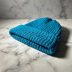Op de afbeelding: Een turquoise gebreide muts met een omgeslagen rand. De muts is gemaakt van een geribbelde gebreide stof. Het ligt op een wit en grijs marmeren oppervlak.