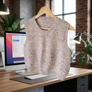 Op de afbeelding: Een lichtbruin gebreid vest met een gespikkeld patroon hangt aan een houten hanger. Het mouwloze kledingstuk heeft een geribbelde zoom en halslijn. De achtergrond omvat een bureau, computer en een bakstenen muur.