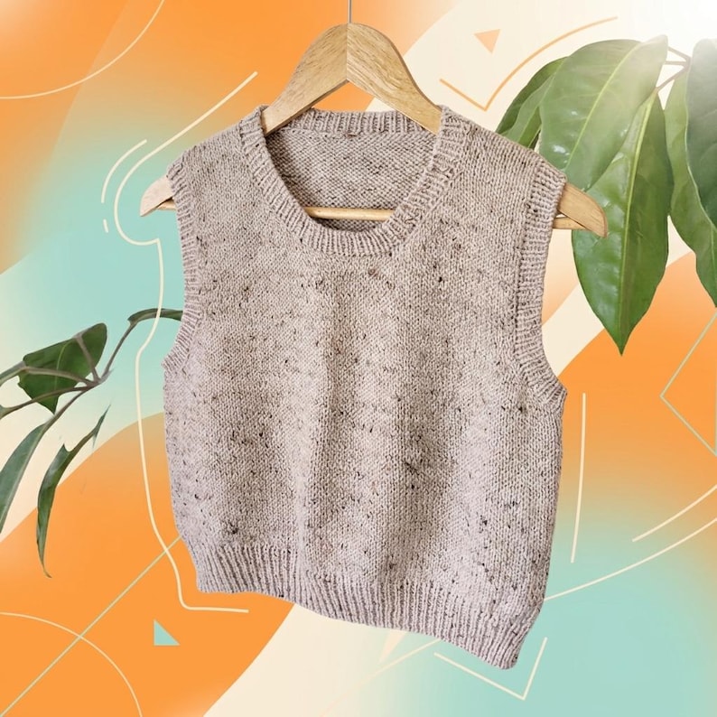 Op de afbeelding: Een beige gebreide vest met een gespikkeld patroon hangt aan een houten hanger. De mouwloze top heeft een geribbelde zoom en halslijn. De achtergrond heeft een verloop van oranje en blauwgroen met groen blad.