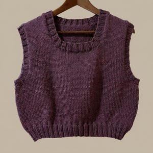 May include: A sleeveless, knitted vest in a deep purple hue. The vest features a square neckline with a ruffled edge and a ribbed hem. The garment is displayed on a wooden hanger against a neutral background.