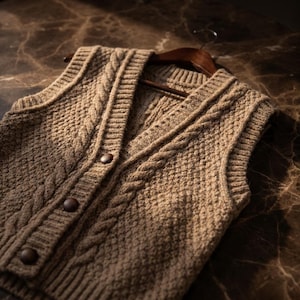 May include: A brown, cable-knit vest with a V-neck and three dark brown buttons. The vest is sleeveless and hangs on a wooden hanger. The knit pattern includes a textured stitch and a cable design.
