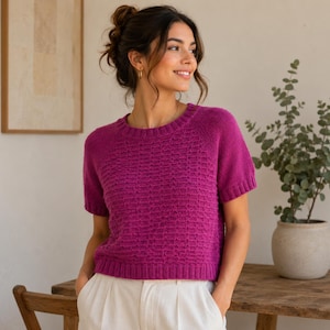 May include: A short-sleeved, magenta-colored knit sweater featuring a textured, repeating grid pattern across the front. The garment has a round neckline and ribbed detailing along the collar, sleeve cuffs, and the bottom hemline of the sweater.