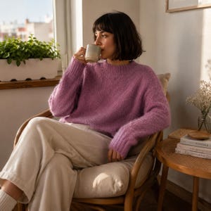 May include: A long-sleeved, fuzzy, mauve-colored knit sweater with a crew neckline and ribbed cuffs, paired with light-colored trousers. The sweater features a soft, textured appearance and a relaxed fit, providing a comfortable look for casual wear.