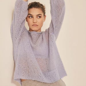 May include: A pale lilac open-knit sweater with long sleeves and a round neckline. The sweater has a relaxed fit. The model is wearing the sweater with neutral-coloured trousers. The sweater is made of a soft, fuzzy material.
