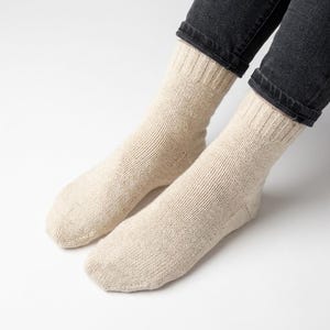 May include: A pair of mid-calf length socks in a light beige, speckled oatmeal color. The socks feature a ribbed knit texture along the cuffs and a smooth, plain knit construction throughout the foot and ankle area, ending at the dark denim jeans.