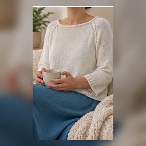 Puede incluir: Un suéter de punto color crema de manga larga con un amplio escote barco, mangas raglán con delicados detalles de ojales a lo largo de las costuras y puños acanalados. El suéter se combina con una falda azul y una manta color crema texturizada.