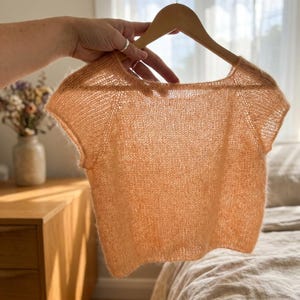 May include: A short-sleeved, peach-colored knit top featuring a wide, rounded neckline and a semi-sheer, loose-stitch construction. The garment is displayed on a wooden hanger, showcasing a lightweight, textured fabric with a soft, fuzzy appearance.