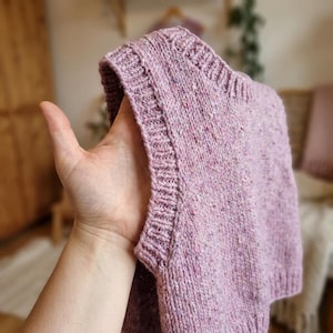 Könnte beinhalten: Ein handgestrickter, ärmelloser Pullover in einem weichen, gesprenkelten Lavendelton. Der Pullover hat einen gerippten Halsausschnitt und Armausschnitte mit einem strukturierten Strickmuster am ganzen Körper. Das Garn hat blaue, rosa und weiße Flecken.